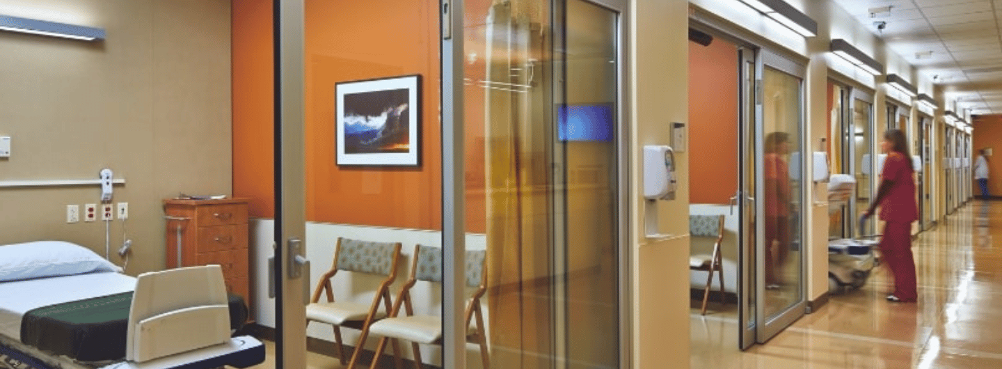 A hospital hallway with glass-walled patient rooms, an empty hospital bed, chairs, and two people in scrubs walking in the background highlights the importance of reliable automatic door service for smooth access and safety.
